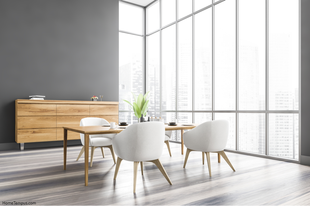 Grey dining room with wooden table and four chairs, wooden chest of drawers, side view. Grey walls and grey wood flooring