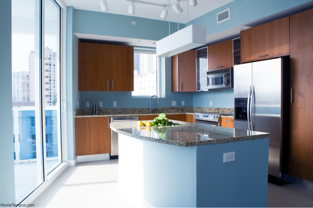 Modern kitchen interior with island in a condo apartment. Brightly lit, light blue walls, granite counter tops, stainless steel appliances.