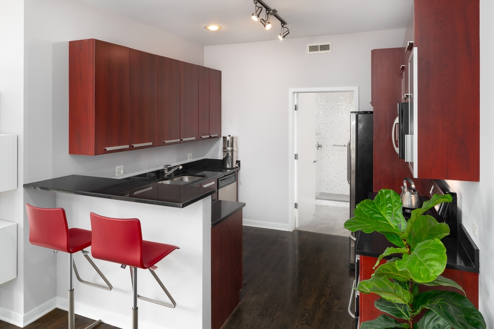 A kitchen with cherry red cabinets, red chairs sitting at a black granite countertop, and dark hardwood floors looking towards a white bathroom.
